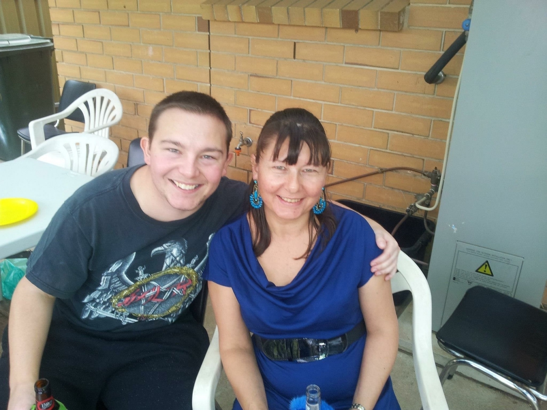 Mother and son sit together on white plastic chairs in an outdoor setting.
